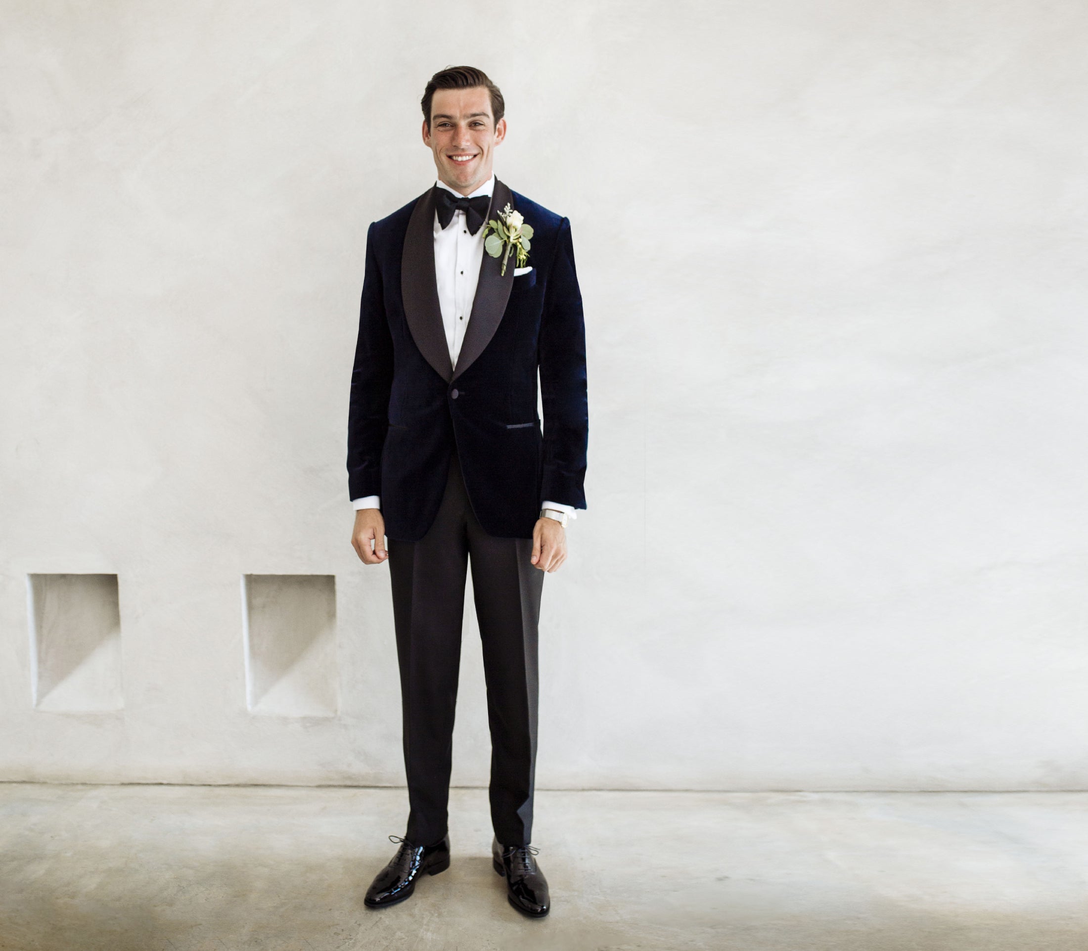 Man wears velvet custom made dinner jacket with curved lapels, white shirt, black bowtie and black trousers. wedding