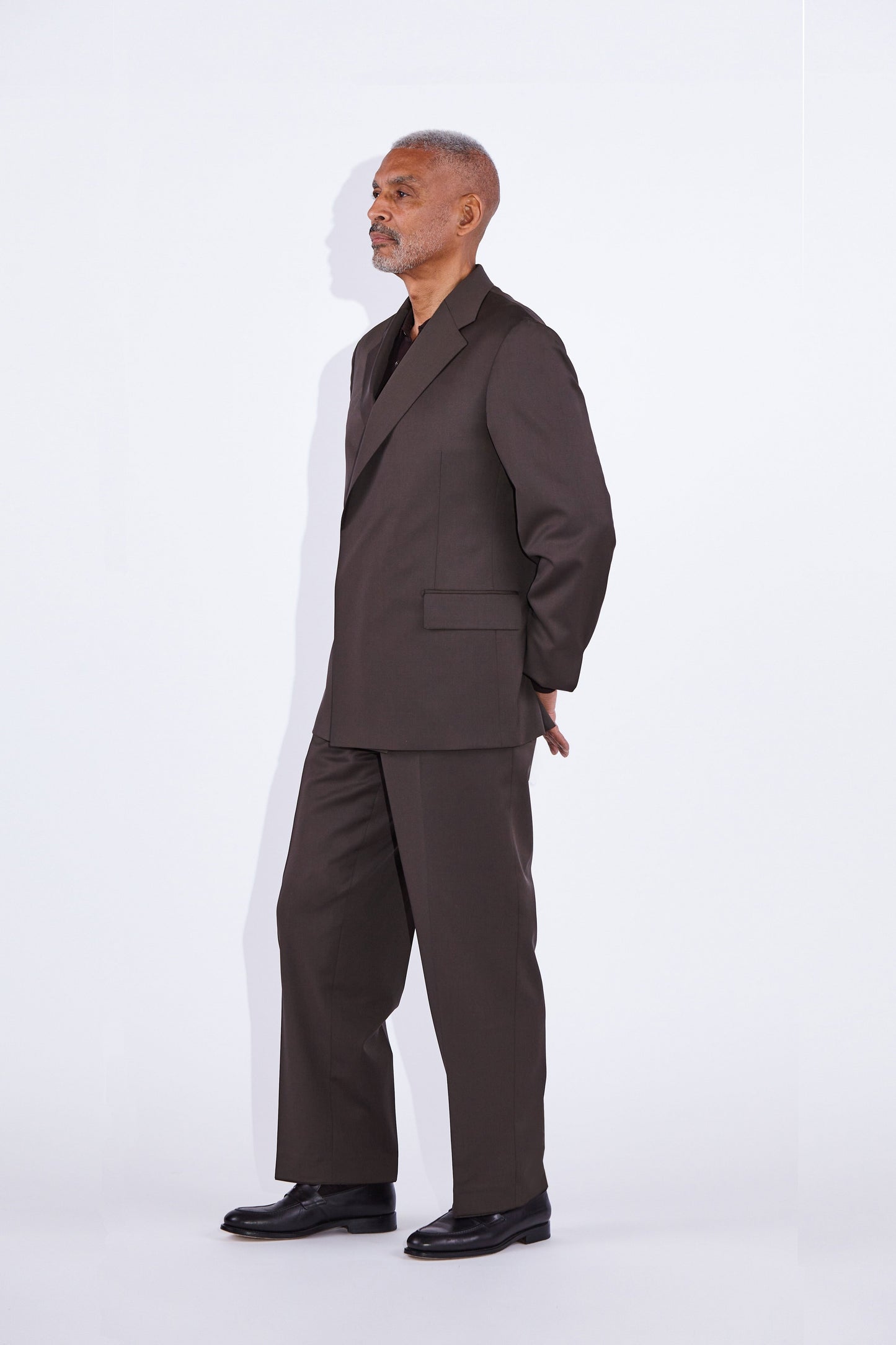 man stands with arms behind his back  notch lapel  double-breasted concealed button brown suit with brown shirt underneath and black loafers