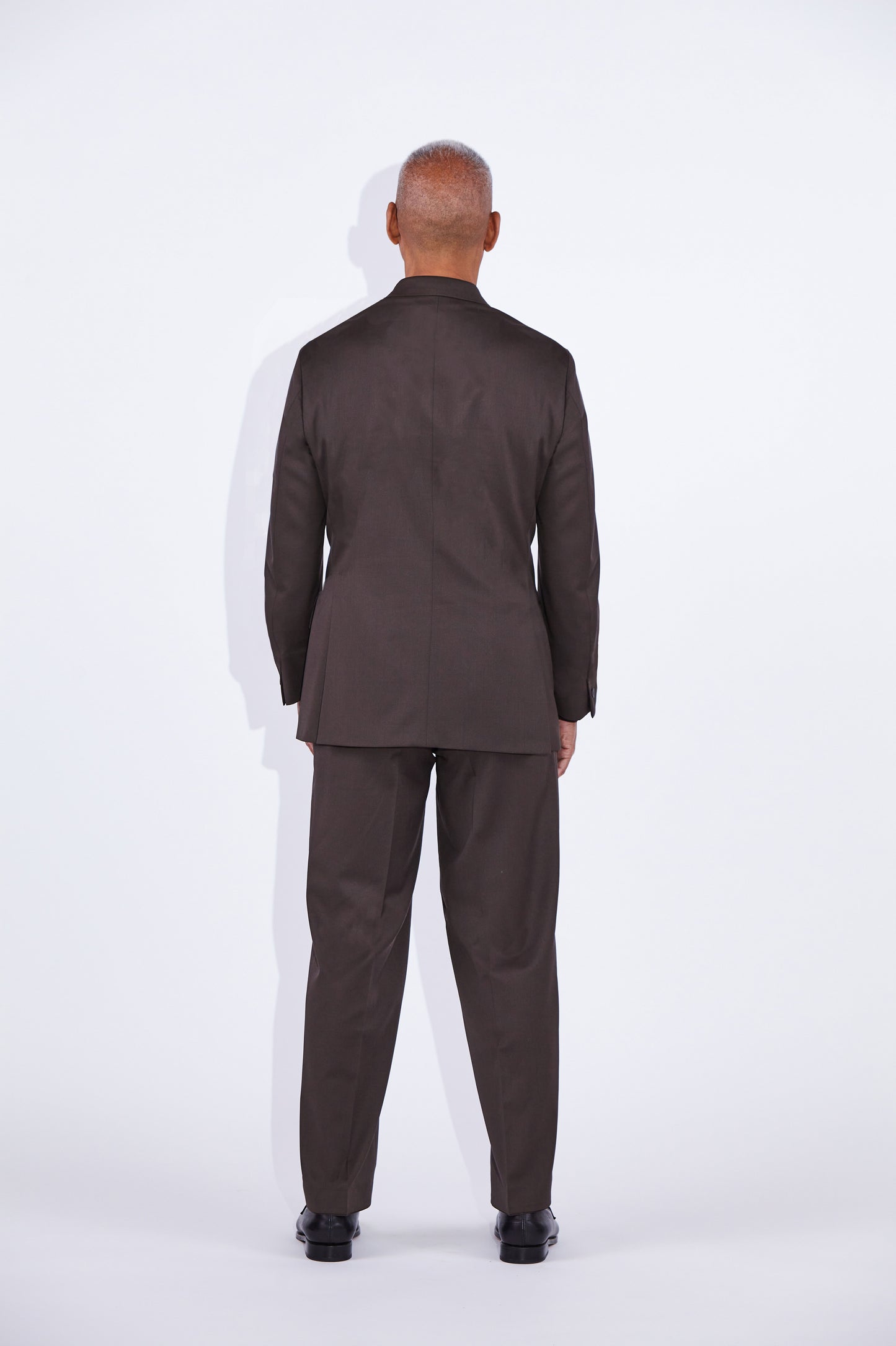 man stands looking away from camera with arms by his side in a   notch lapel  double-breasted concealed button brown suit with brown shirt underneath and black loafers