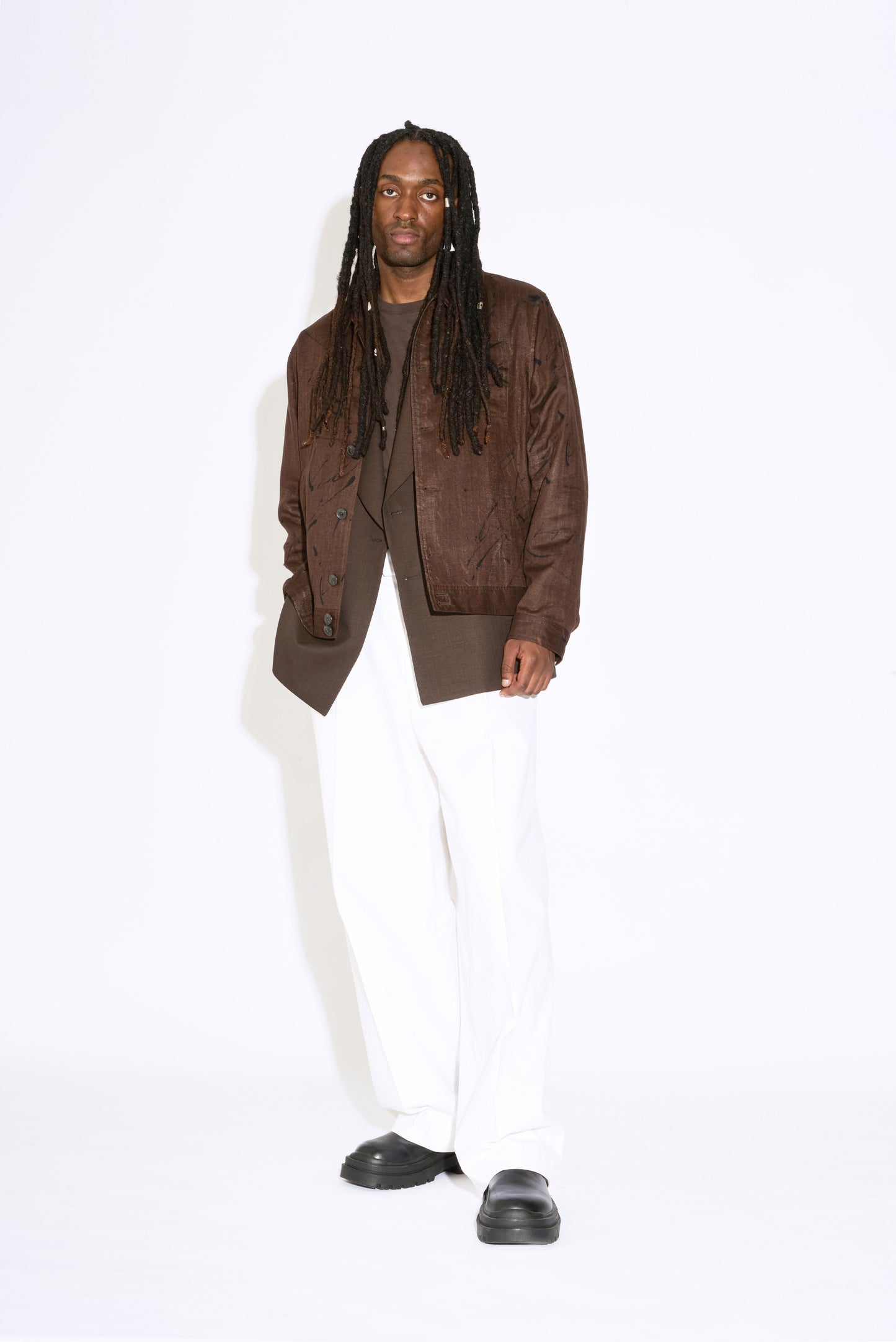 man with dreadlocks stands wearing brown bomber jacket and white wide leg trousers with black chunky shoes