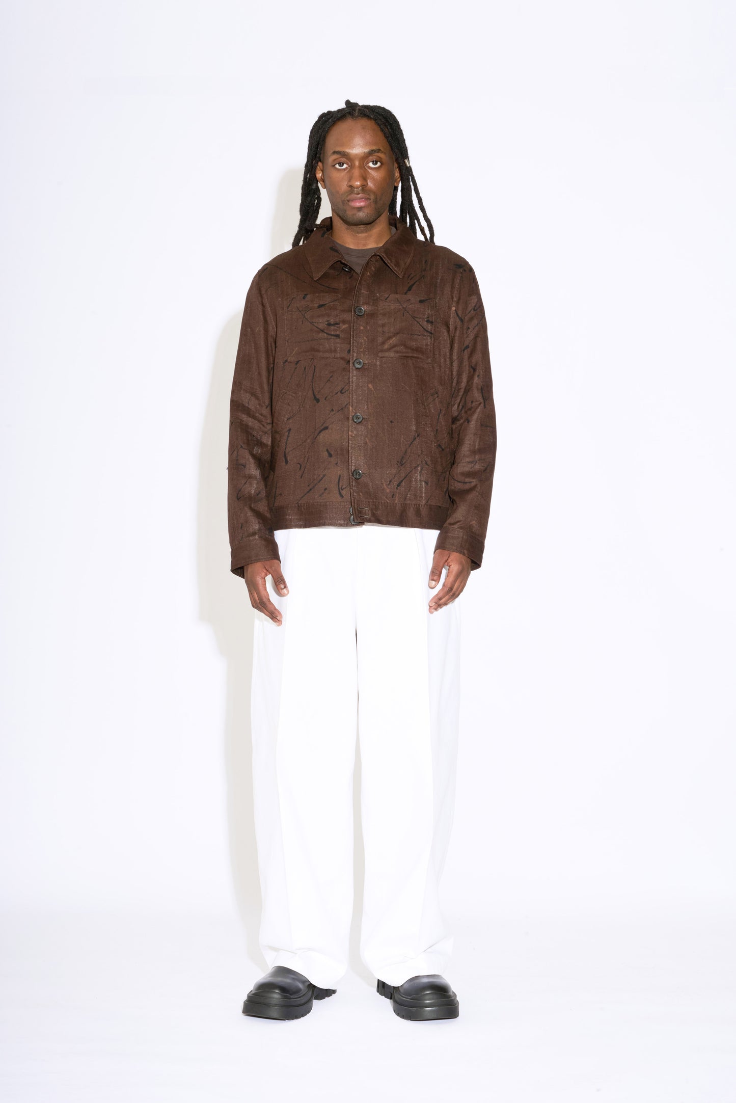 man with dreadlocks stands straight wearing brown bomber jacket and white wide leg trousers with black chunky shoes