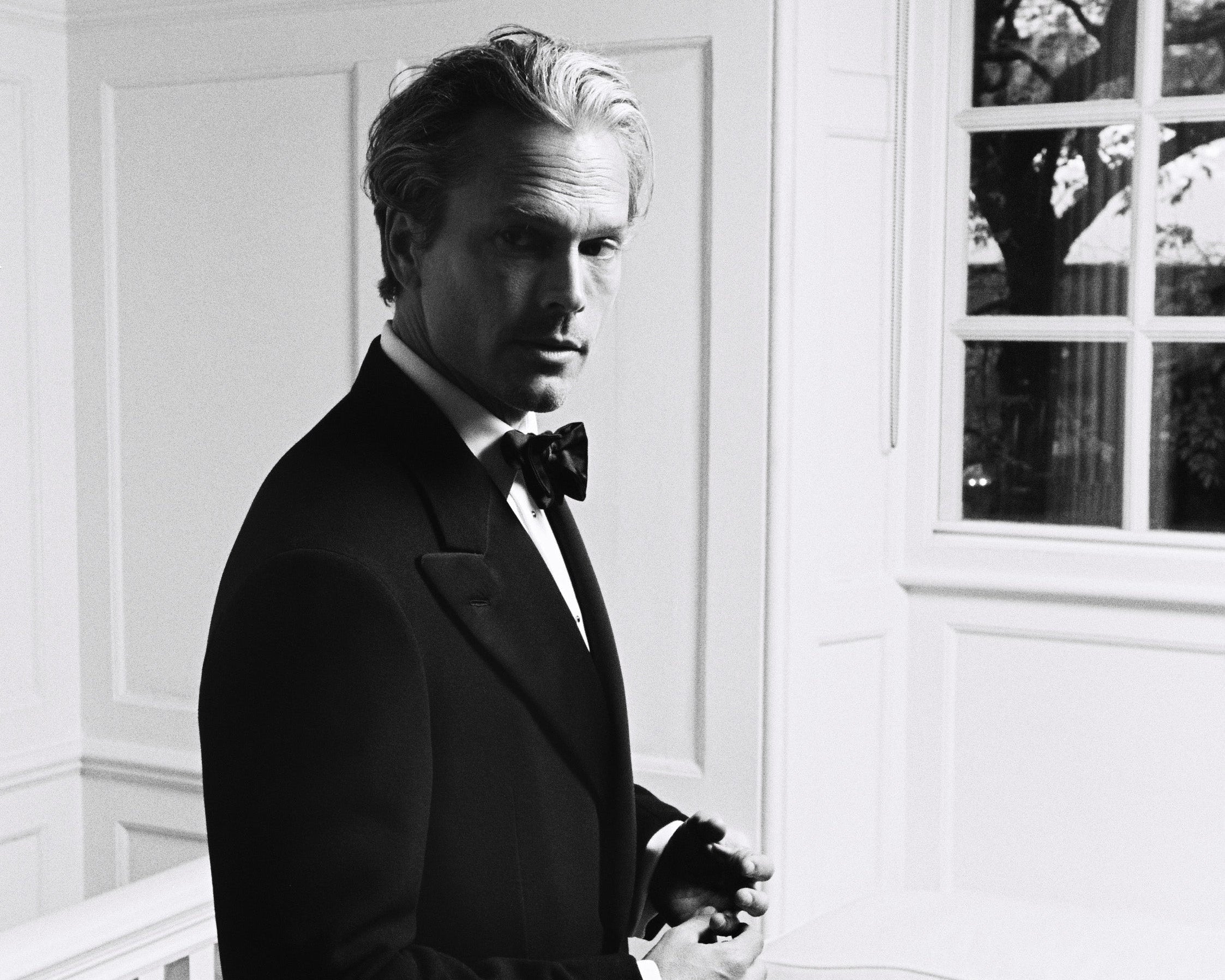 Man in a tuxedo standing in a room with white walls and a window.