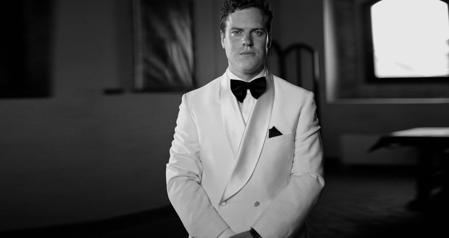 Man wears custom made 2 button white dinner jacket, with white shirt and black bowtie. Worn for his wedding. Black and white image