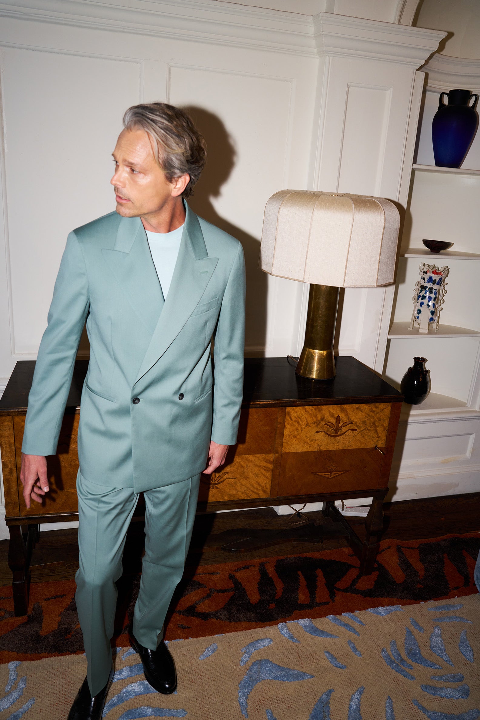 Model in a teal Casely-Hayford suit standing in a room with a lamp and decorative items.