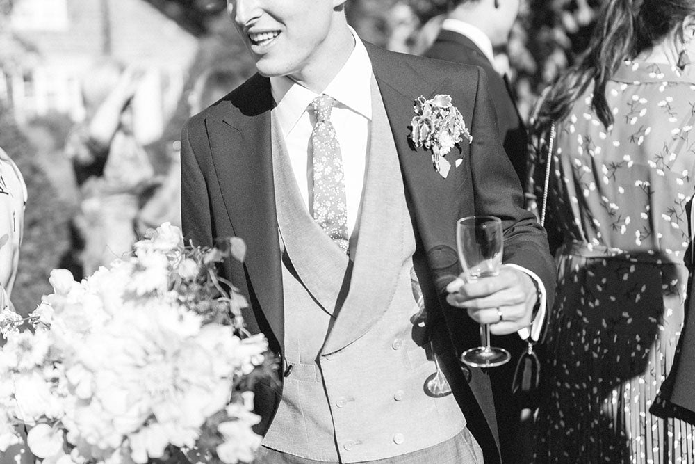 Close up of a man on his wedding day wearing a custom made blazer with a 6 button waist coat and shite shirt with a floral tie. Black and white image 