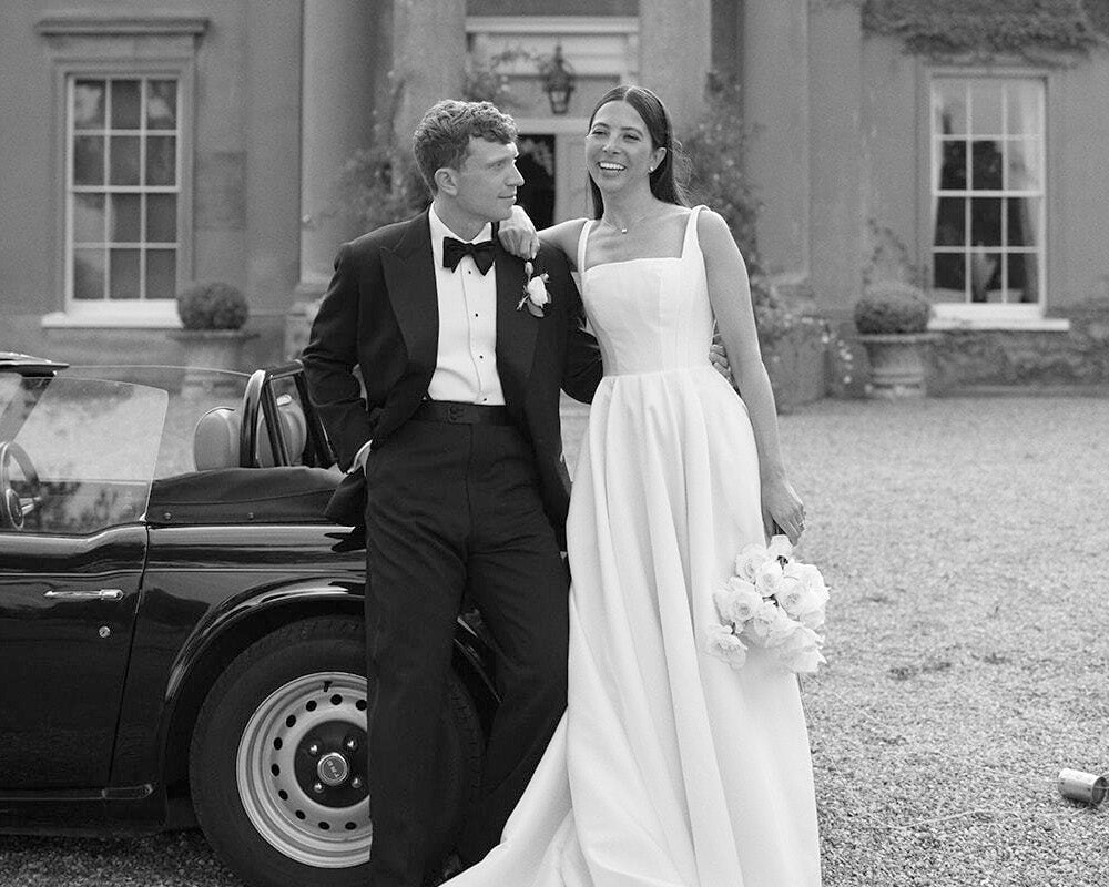A black and white photograph of a bride and groom standing outdoors in front of a grand classical building. The groom wears a tuxedo and stands next to a vintage convertible car, while the bride wears a sleeveless white wedding gown and holds a bouquet. Groom wearing Casely-Hayford.
