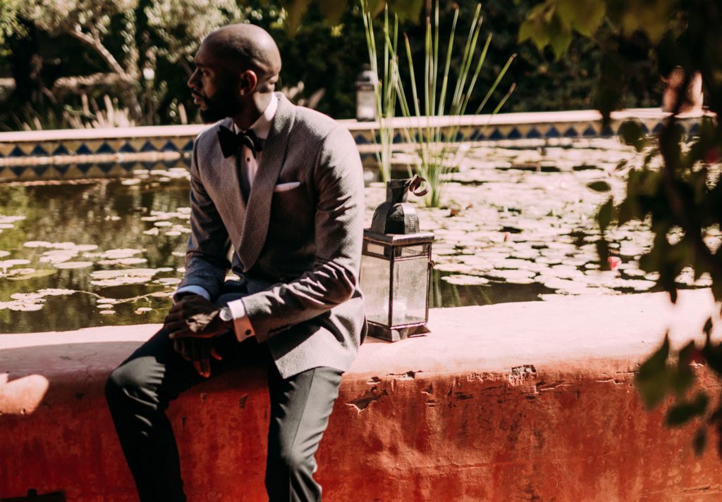 Groom in grey dinner jacket , white shirt and black bow tie and black trousers, sitting on a red wall by a pond