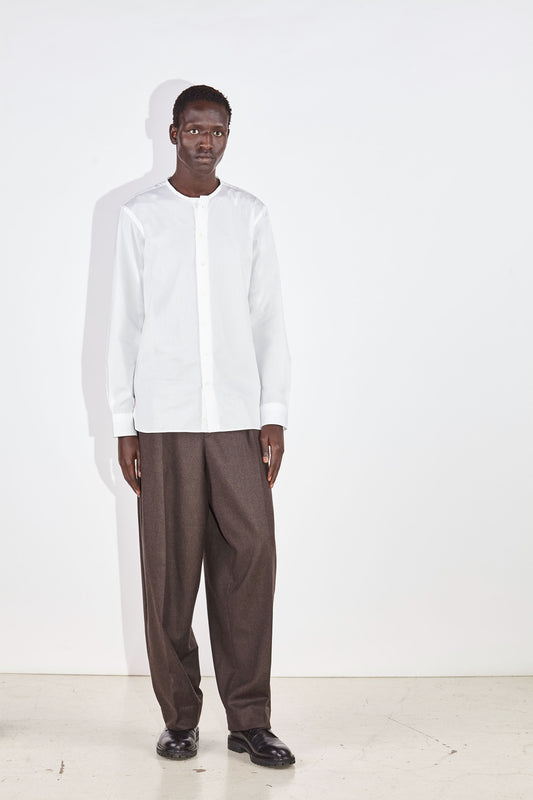 a man standing straight in a collarless white shirt and wide brown trousers 